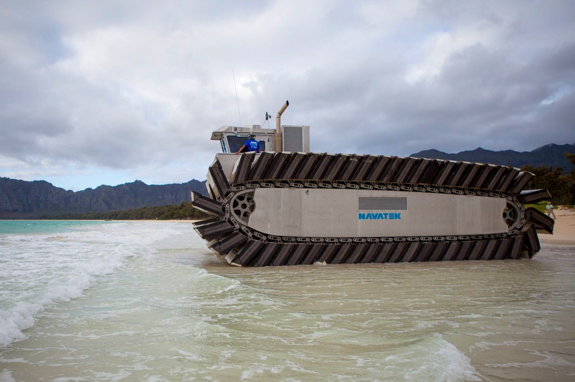 A futuristic large tank-like vehicle comes out of the water onto a beach under a cloudy sky and against a background of rocky cliffs. A single individual rides on top of the vehicle.