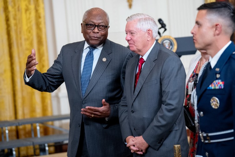 Two political figures visit during an event at the White House. The individual on the left gestures while speaking, while the other individual listens and smiles. On the far right of the image, there is an out-of-focus soldier in a formal uniform.