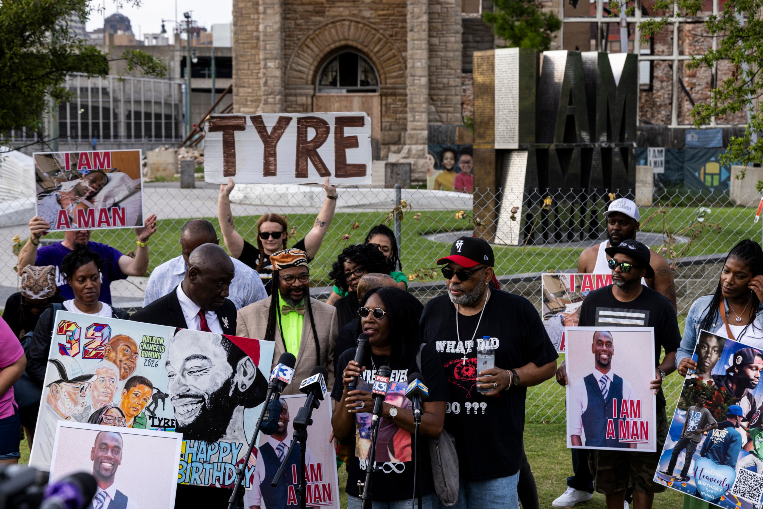 A woman speaks into numerous microphones. She is in the midst of a crowd of people outdoors. The other people hold signs that say, “Tyre” and “I am a man.” Behind them, a large sculpture spells out the words “I am a man.”
