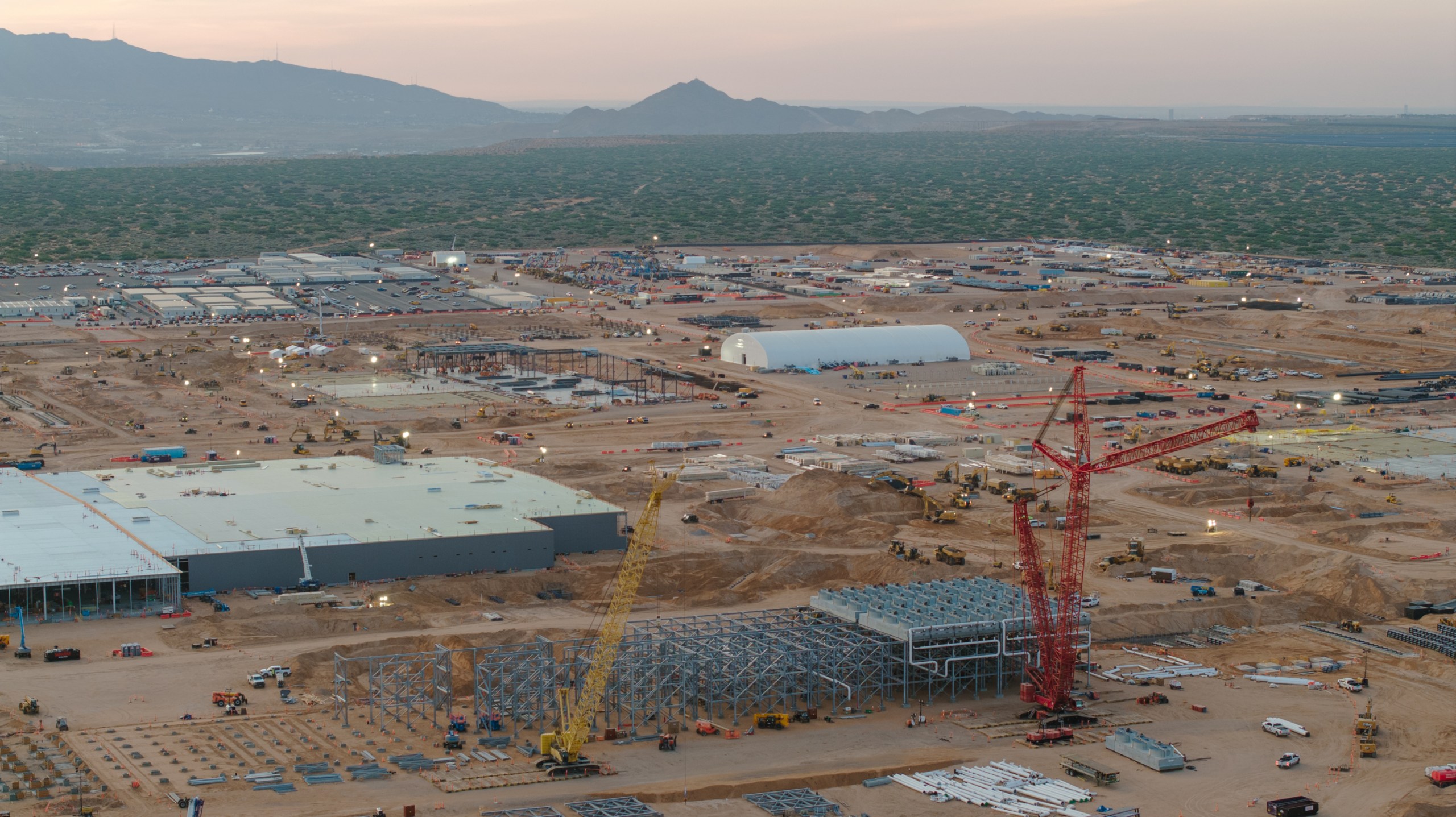 An elevated view captures a sprawling construction site filled with large steel structures, heavy machinery and prominent yellow and red cranes set against a mountain backdrop at dusk.