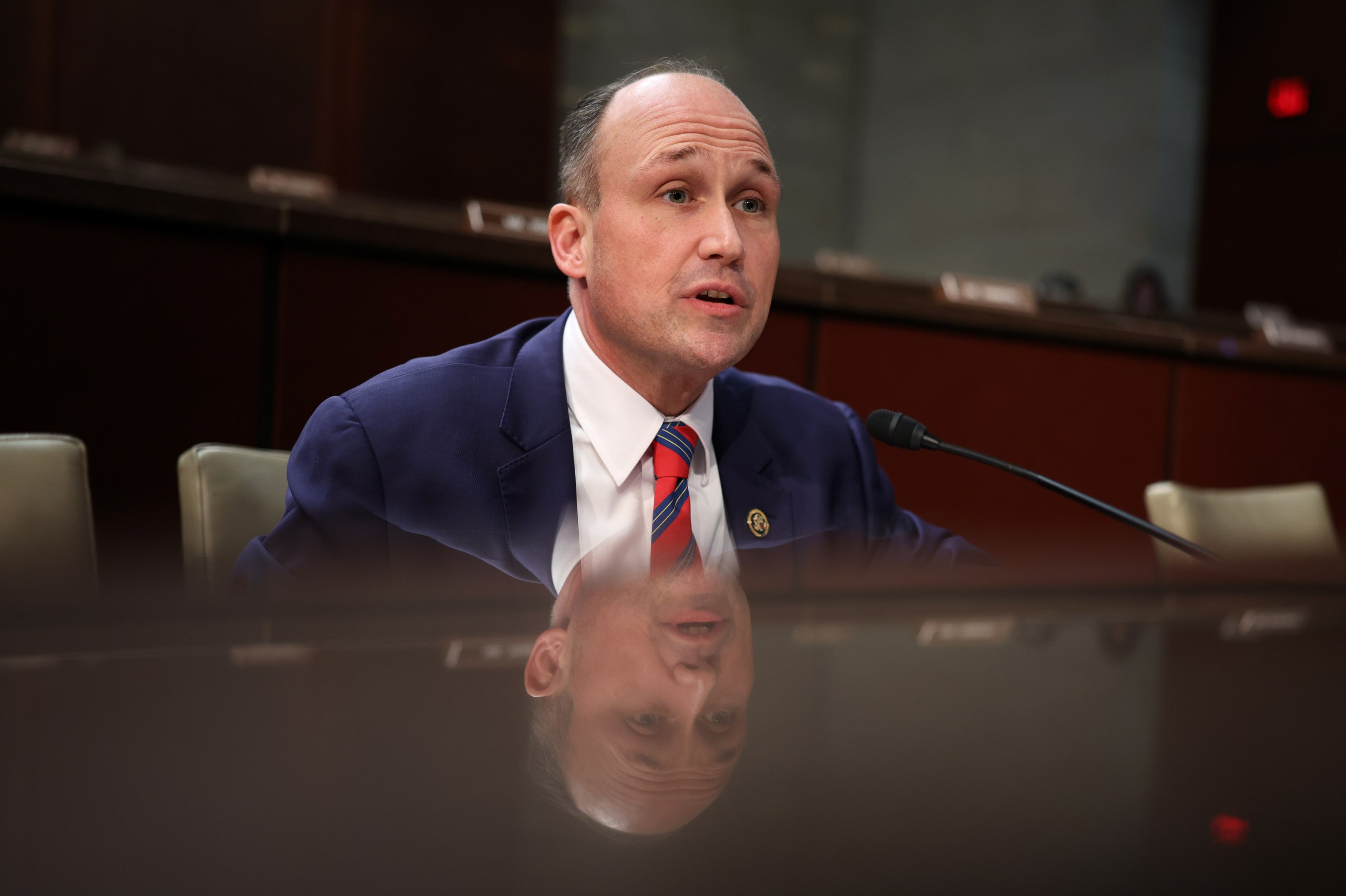 A man wearing a suit and tie sits at a wooden table with a microphone. His reflection can be seen in the table under him.