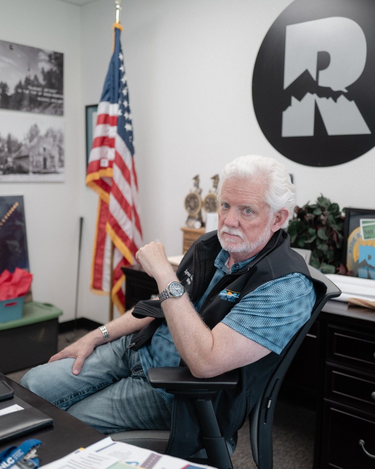 A man with white hair and a white beard sits at an office desk in front of an American flag and village logo, resting his arm on his chair while looking toward the camera.