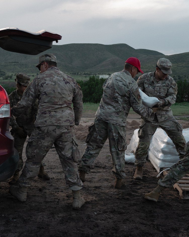National Guard service members load white sandbags from wooden pallets into the open trunk of an SUV ahead of flood preparations.