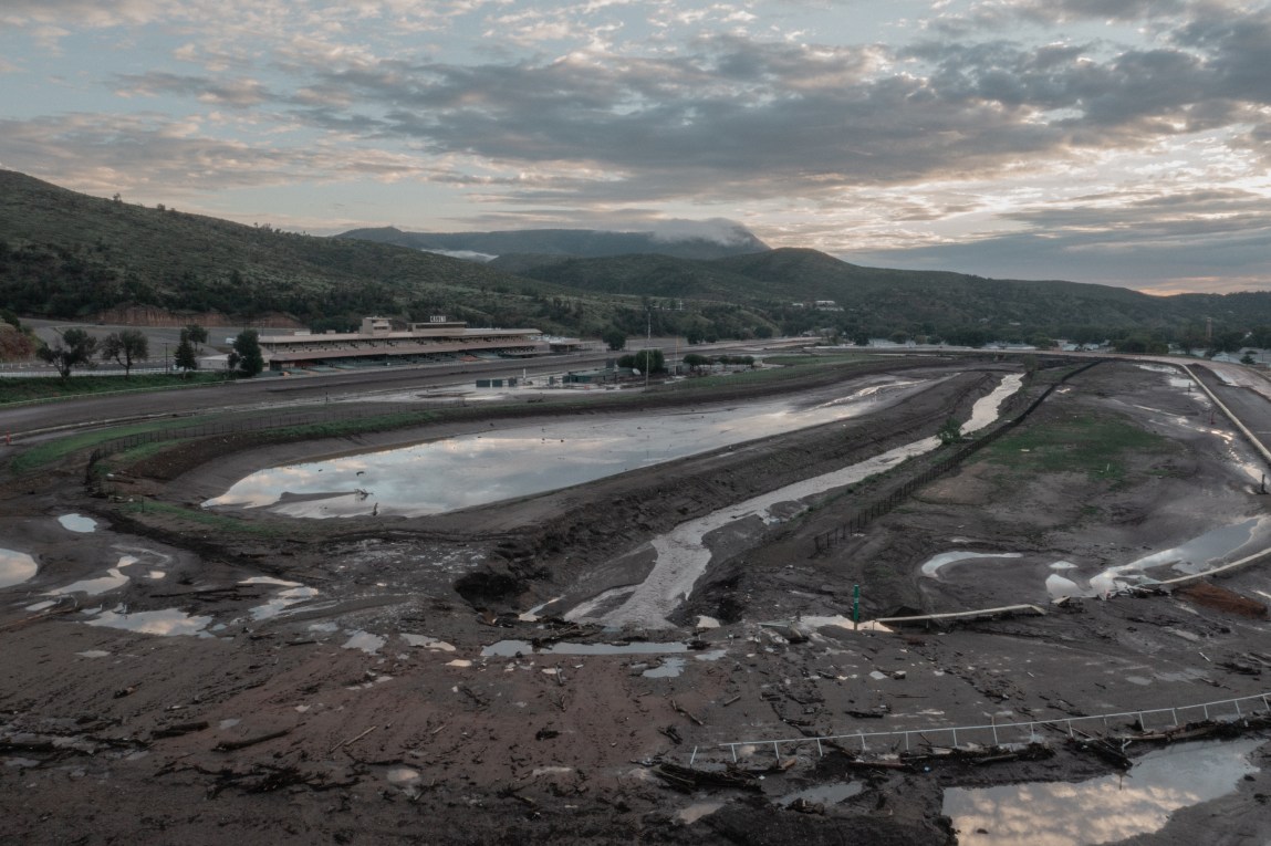 An elevated view shows a mud-caked, flooded horse-racing track surrounded by standing water and mountain ridges under a cloudy sky.