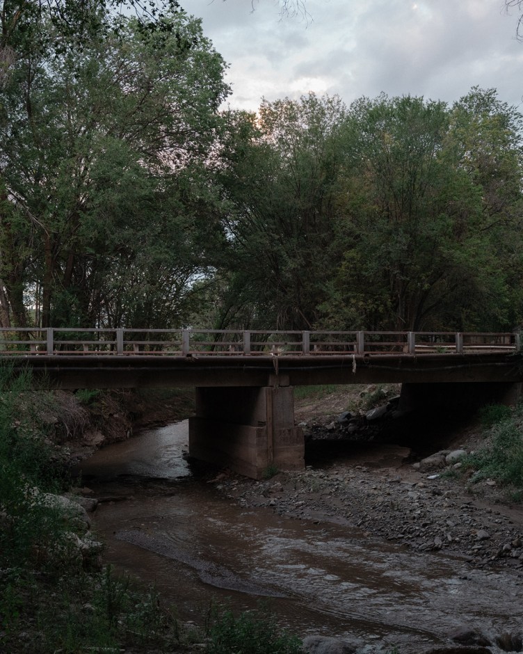 A narrow, concrete beam bridge spans a shallow, muddy stream flanked by dense green trees and dirt banks.