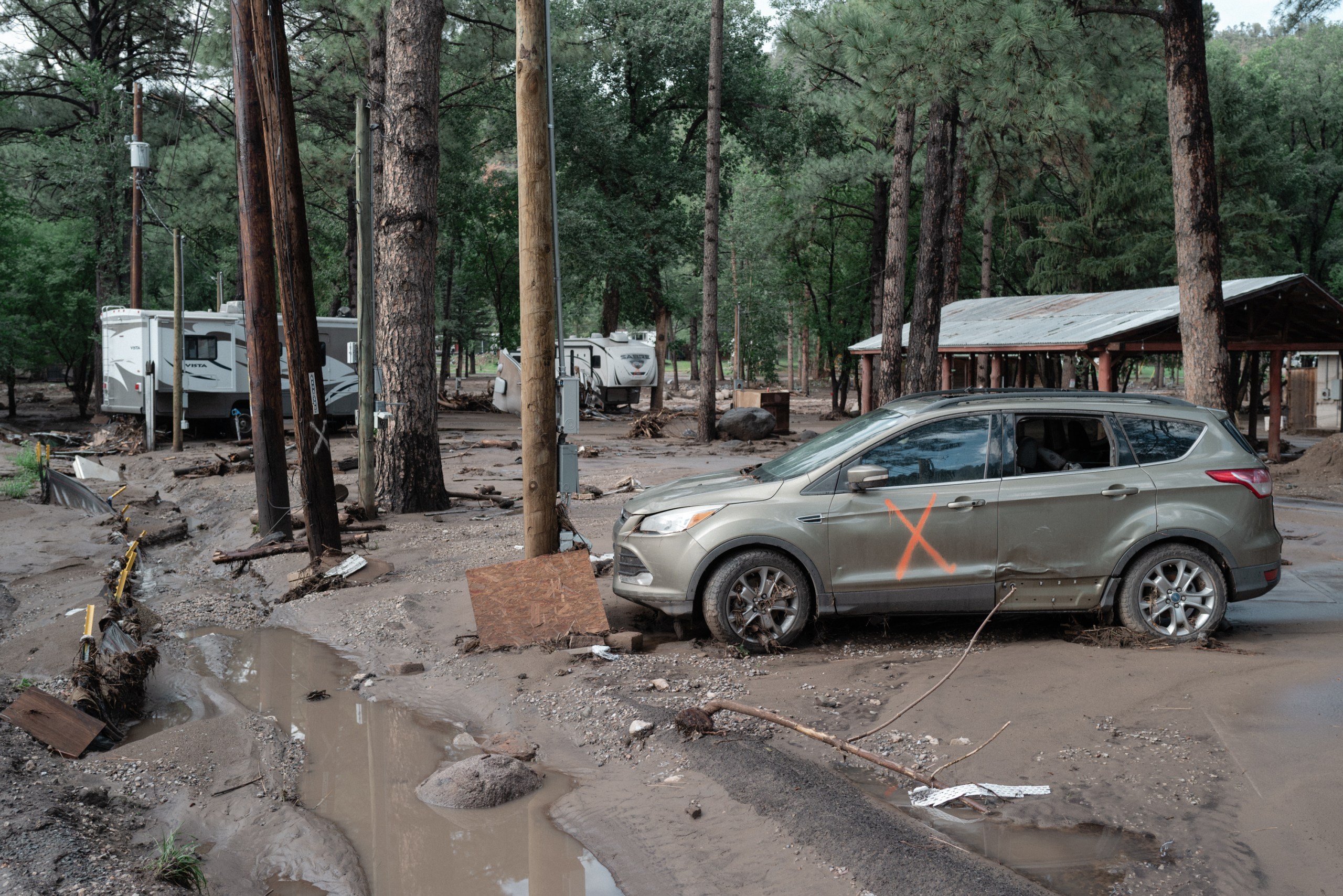 An SUV marked with an orange X and RV trailers sit stranded in mud and debris following severe flash flooding in a wooded area.