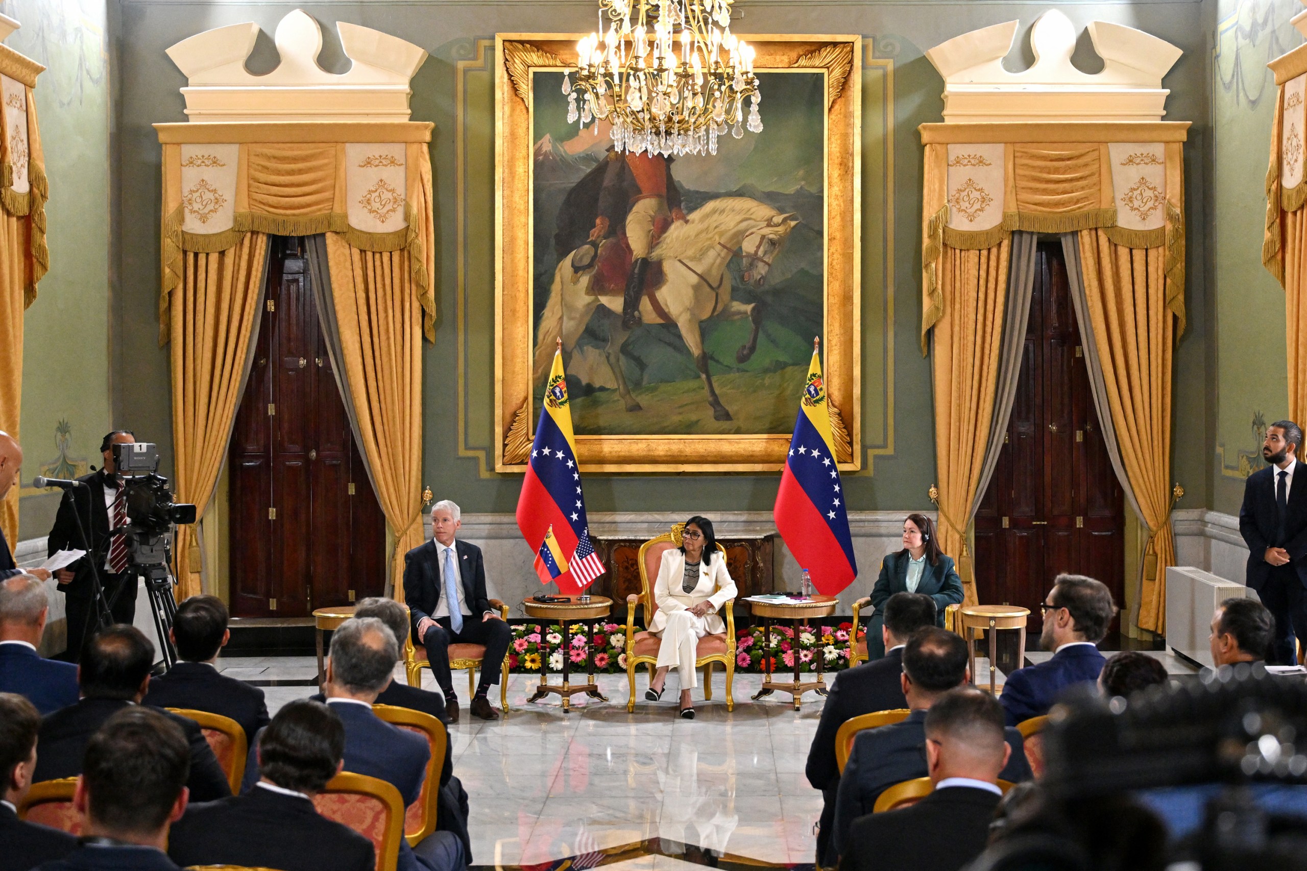 A woman in a white suit sits in a large room in front of a crowd of people. A man in a suit sits to her right, and a woman in a suit sits to her left. She is flanked by two Venezuelan flags. The three of them look toward TV cameras off to one side. The room is lavishly decorated, with a chandelier, long golden curtains over the doorways and a large framed painting of a person riding a horse.