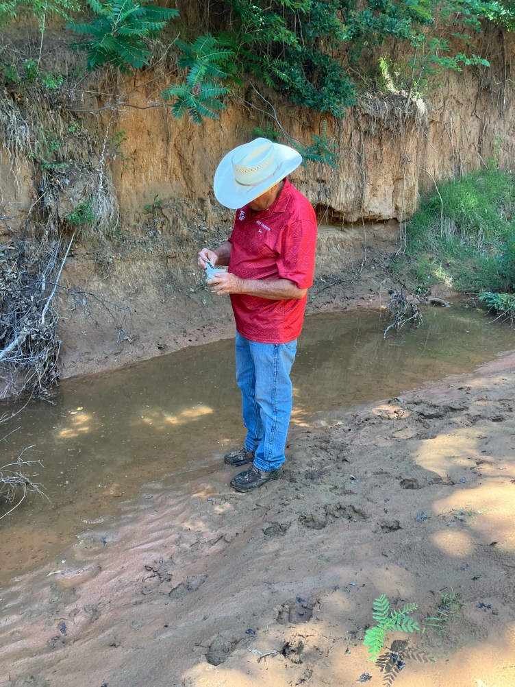 A man in a red shirt and cowboy hat holds a small device with its end in a cup while he stands on muddy ground.