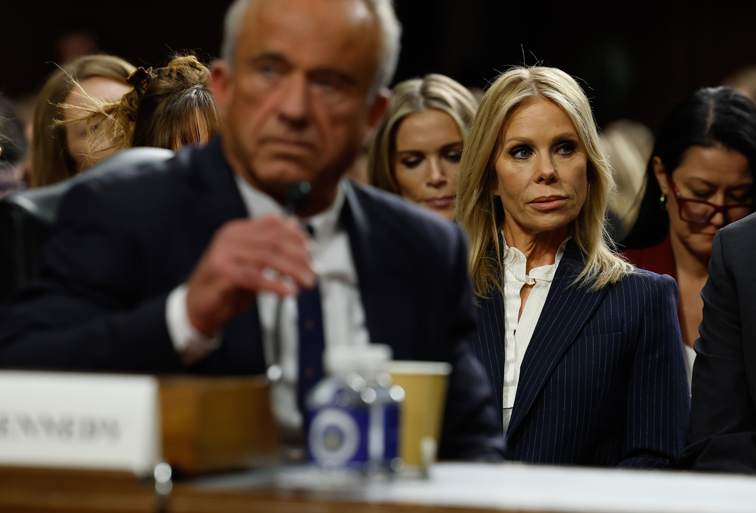 An out-of-focus man in a suit and tie sits at a table in the foreground while adjusting a microphone during a Senate hearing. A woman in a business suit is in focus just behind him and is surrounded by many people.