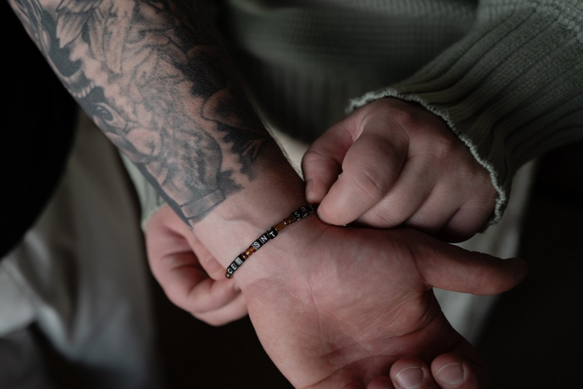 A close-up shot of hands touching a beaded bracelet worn on a wrist below a tattooed arm.