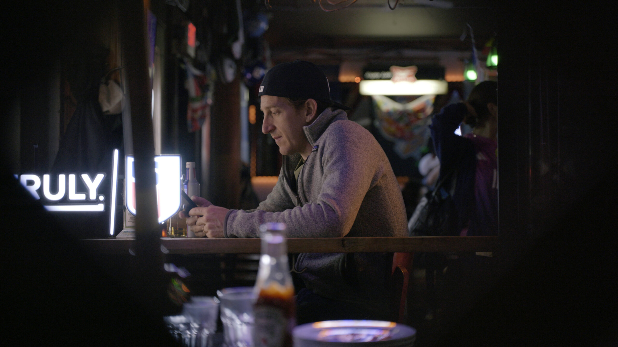A man wearing a backward baseball cap sits in a dark bar, staring at his phone.